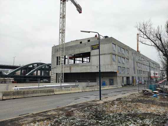 A road runs to the right of the new building. All window openings are provisionally covered with plastic sheets (protection against cold). A crane stands in front of the building, the railroad bridge can be seen on the left. Cloudy and cold weather just above freezing point.