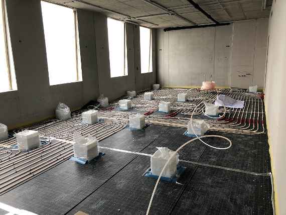 Blick in einen Raum. Auf dem Boden sieht man die teils verlegten Kunststoffschläuche der künftigen Fußbodenheizung, die festmontiert sind. Unter den Schläuchen liegen Isoliermatten.