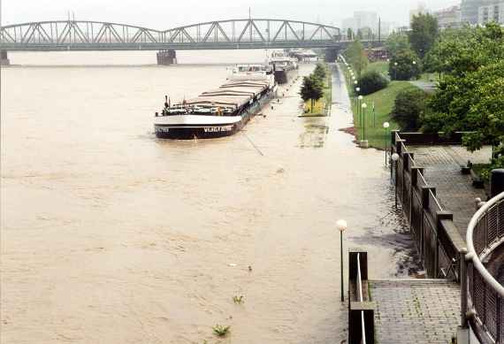 Hochwasser an der Donau, im Hintergrund sind eine Brücke und mehrere angelegte Lastschiffe sichtbar, der Gehweg entlang der Donau ist überschwemmt.