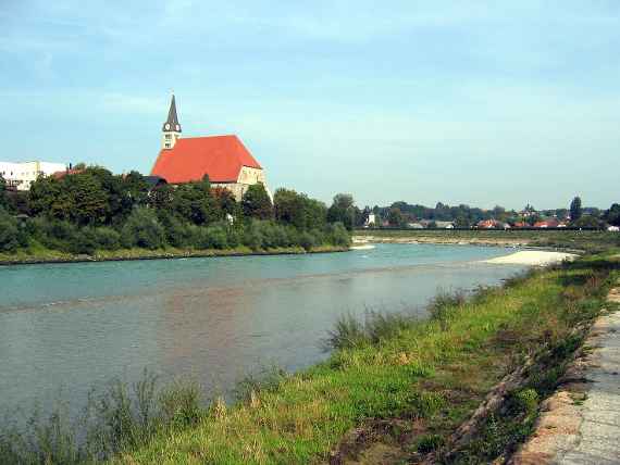 Bild des Flusses und der mit Gras und Büschen bewachsenen Böschungen. Im Hintergrund sieht man den Turm und das rote Ziegeldach der Kirche von Laufen.