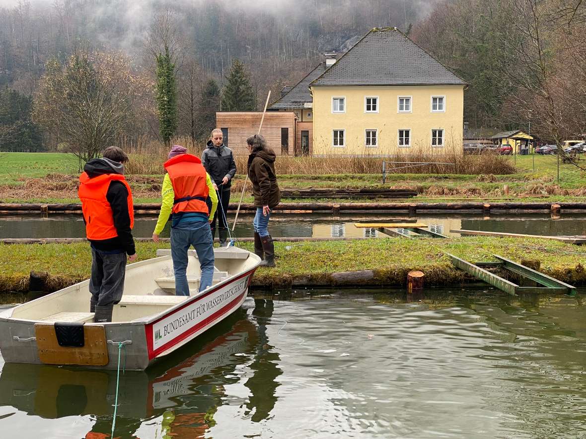 Die Teilnehmenden befinden sich bei einem Teich. 2 Personen sind einem Boot und 2 zwei Personen befinden sich auf der Wiese neben den Teich. Den Teilnehmern wird hier die Vermessung der Anlage beigebracht.