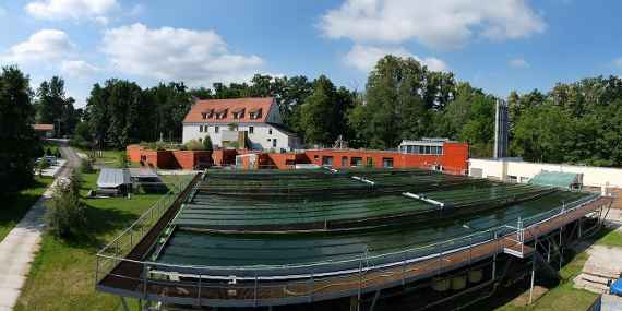 The picture shows the Algatech Centrum in Trebon in the Czech Republic. In front is a large fish farming facility, behind it is a house, further back is forest area. Taken in sunny and cloudy weather.