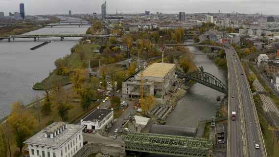 View of the new building, on which the new wooden roof beams can be seen. In front of it the Schemerl bridge and the Nussdorf power plant. A four-lane road runs to the right (B14). Behind the new building, a railroad line runs across the Danube Canal. At the rivers left the Danube and right Danube Canal are to be seen, behind it the 20th district. Far back on the left is the DC Tower skyscraper.