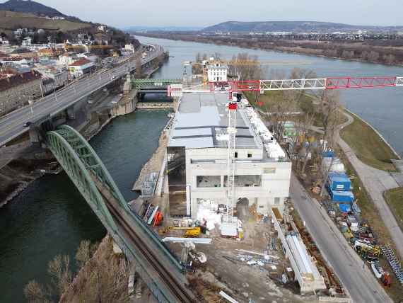 View from above at the rear entrance. Most of the roof is covered with bitumen sheeting. In front of and behind it a construction crane. On the left of the building, one can already see the exterior façade, which is mirrored from the bottom up. Far left the Danube Canal, a crossed railroad bridge and almost parallel running highway, behind it houses (Nussdorf). Far left above is the (hilly) Kahlenberg. Behind the new building is the building of the Magistrate's Department of Viennese Waters, to the left of it over Danube Canal the Schemerl Bridge. On the right the Danube, behind it direction Langenzersdorf (Lower Austria). A road leads past the building on the right in the direction of the Schemerlbrücke. Various construction site equipment and materials on both sides of the road.