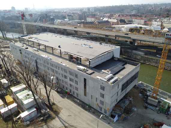 Photo taken obliquely by drone. On the right you can see the future main entrance. Almost all windows are covered with tarpaulins. On the roof you can still see some scaffolding and building materials. Behind the house runs the Danube Canal, running parallel is the highway. Behind it is the 20th district, sunny and dry weather.