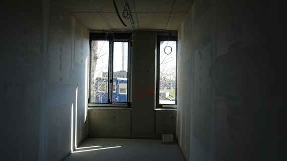 Picture of a room in the building from the door side. Towards the window, you can see the two windows already installed. At the top, the power cables for ceiling lights run, some hang rolled up down at the window area. Left and right are already filled plaster walls. Right in front of the window, you can see a shaft for future power and EDV supply of this room.