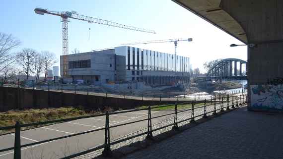 Oblique view to the new building, taken below the highway, in front of it runs the Danube Canal. Two construction cranes can be seen in front of and behind the new building. The outer facade is taking shape. On the far right the railroad bridge can be seen in sunny and dry weather.