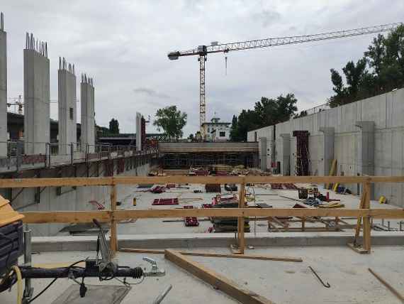 A view of the interior from the first floor. On the left and right raised walls. In the middle are building materials (wood, formwork and so on). In the very front you can see the crane (one of two, the other is behind the photo), further ahead you can see the Nussdorf power plant and the Schemerl bridge. Dry pleasant weather, no sun.