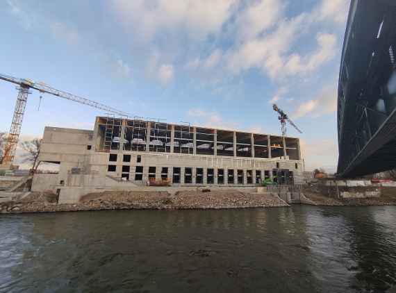 View over the Danube Canal, construction cranes are on the left and right. The roof is "closed" and you can see the concrete steps on the left side of the building, on the right side the first cladding has already been installed. Two construction vehicles in front of the building. Dry, cold weather and a bit cloudy. On the far right the railroad bridge over the Danube Canal.