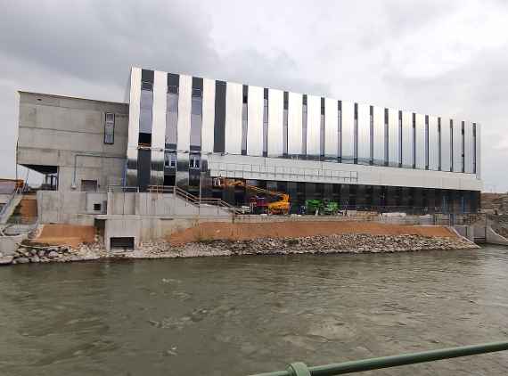 View over Danube Canal. All windows already installed and 80% clad all around, which wink silver due to the weather. On the left side you can see the future main entrance. Two lifting platforms with construction workers can be seen in front of the building. The embankments on the bank of the Danube Canal are covered with geotextile. Dry and cloudy weather.