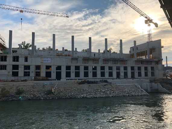 Concrete columns for the walls of the future test hall rise into the sky; a construction crane stands on each side. The Danube Canal flows in front of the building.