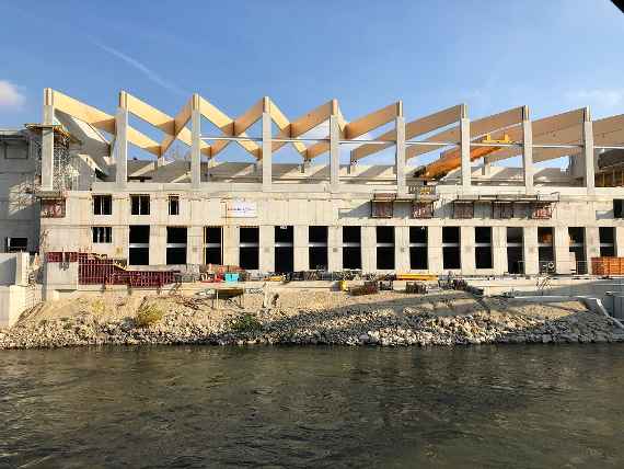 The shell construction is almost finished. About 25-metre-long wooden beams have been mounted as supports for the roof. The Danube Canal flows in front of the building. The sun is shining on the building.
