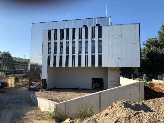 A view from the rear side of the building, where there will be some parking spaces in the future, including access for trucks to the building. All areas are still unpaved. On the right is the access road to the test hall, currently still without a gate. Left on photo in front of the building a small excavator, further left the railroad bridge, behind it the highway. The photo was taken in sunny and warm weather.