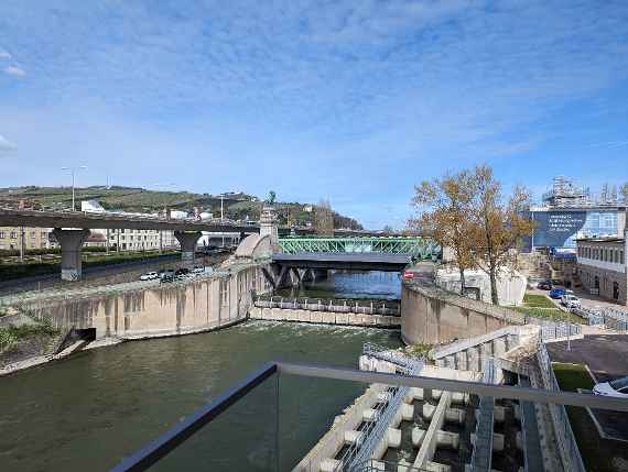 Oblique view from the main building on the right. Visible is the Nussdorf weir, behind it the Schemerl bridge. Running obliquely to the left are the highways stilts, behind them are some residential buildings from the nineteenth district and further back the Kahlenberg. Right in front the fish ladder, our visitor parking lots, and just behind it the building belonging to viadonau. Further behind, the building of the Vienna Waters (Magistrate's Department 45), currently scaffolded for renovation.