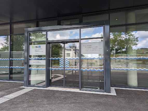 The picture shows the main entrance with a large sliding door that opens to the left and right. To the left of the sliding door is the logo of BOKU and the text "IWA Institute of Hydraulic Engineering, Hydraulics and River Research" and to the right is the logo of the Federal Agency for Water Management and below the text "Federal Agency for Water Management, Institute for Hydraulic Engineering and Hydrometry" in German. Below the logos, two parallel, wavy lines run across the entire glass surface, the upper line is white and the lower line is blue. The wavy lines are repeatedly interrupted by a white BOKU logo. Where the two sliding door leaves meet, there are four more blue and white wavy lines in the glass. On the far left you can clearly see a floor guidance system for the blind.