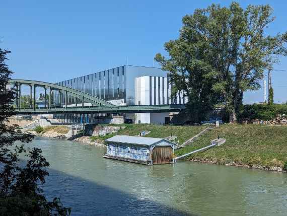 The picture shows the Danube Canal in the foreground and the building of the hydraulic engineering laboratory on the other bank. A boathouse floats in the Danube Canal and is attached to the land with thick steel pipes. A railway bridge runs in front of the hydraulic engineering laboratory. On the right are two large trees in front of the railway bridge. If you look further to the right of the picture, you can see a chimney with grooves; this is where the ship lock between the Danube and the Danube Canal is located. On the left side of the Danube Canal you can see a shadow, this comes from a road bridge running along the Danube Canal. On the far left are some bushes. The picture was taken in sunny and cloudless weather.
