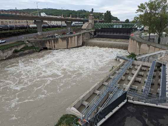 The picture shows the weir on the front side, on the right the fish ladder. The photo was taken on 30 August 2023, after a flood alert had been issued in western Austria in the preceding days due to storms coming from Italy. You can see very turbid water on the Danube Canal. Two segmental weirs are lowered to limit the flow and to collect the debris, mainly wood, from the water surface. On the left the motorway bridge and behind it the Nußdorf railway station.