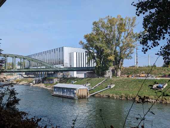 The picture was taken obliquely sideways, from a point of view near the Danube. In a meadow there are some tall trees in front of the building running parallel to the road. In the meadow are two soccer goals, but without nets. Behind them is a fence with access to the canal, through which Danube water can be directed to experimental facilities in the building. The facade of the building is divided into vertical stripes (dark gray and white). In the dark vertical stripes are the windows of the office and laboratory rooms. Three white poles on the roof are lightning rods. The picture was taken in the somewhat sunny afternoon.