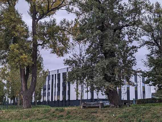 The picture shows the Danube Canal in the foreground and the building of the hydraulic engineering laboratory on the other bank. In the Danube Canal floats a boathouse that is attached to the land with thick steel pipes. A railroad bridge runs in front of the hydraulic engineering laboratory. On the right there are two big trees in front of the railroad bridge. If you look further to the right of the picture, you can see a chimney with grooves, there is the ship lock between the Danube and the Danube Canal. On the left side of the Danube Canal you can see a shadow, this comes from a road bridge running along the Danube Canal. On the far left are some bushes. The picture was taken in cloudy and cloudy weather.