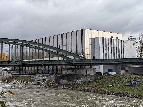 The picture, taken from the south at an angle, shows the Danube Canal in the foreground and the hydraulic engineering laboratory building on the other bank. It is a very light-coloured building. The windows can be recognised as vertical, darker stripes. A railway bridge is visible in front of the building, which runs over the Danube Canal. Some company cars can be seen in the delivery area of the building. The photo was taken in autumnal and cloudy weather.