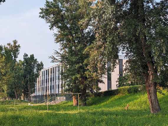The picture was taken from an oblique position, at a viewpoint near the Danube. There are some tall trees on a meadow in front of the parallel to the road erected building. There are two football goals in the meadow, but without nets. Behind is a fence with access to the canal, through which Danube water can be channelled into experimental facilities in the building. The facade of the building is divided into vertical stripes (dark grey and white). In the dark vertical stripes are windows of the office and laboratory rooms. Three white poles on the roof are lightning rods. The picture was taken in the early, sunny morning.