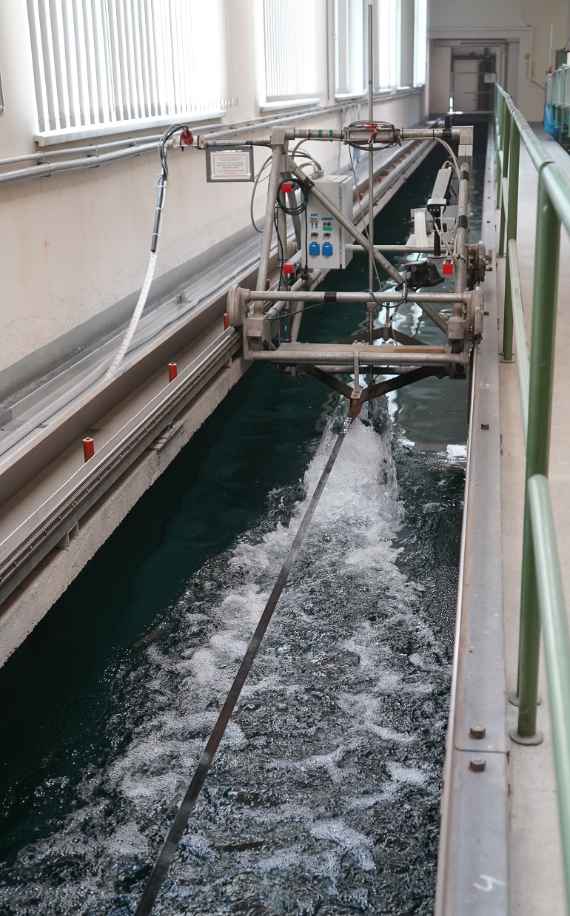 The measuring carriage travels with the current-meter installed in the calibration unit. The water behind it foams due to the body of the meter. On the left some windows with blinds and on the right the railing which protects the employees from the calibration plant.