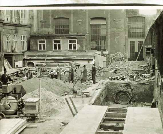 A black and white photo shows a concrete slab and a pit on the right. On the left is a concrete mixer and scattered on the site five construction workers with various building materials.