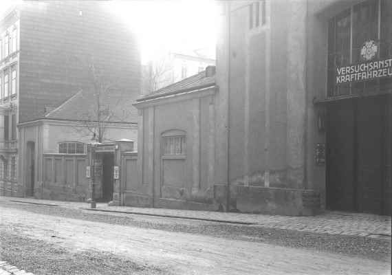This picture shows in black and white how it looked at that time. On the far left is the residential house with 4 floors, which still stands today, in front of it is the garage entrance (high entrance at that time), the window of it shows the actual room of the workshop. In the middle the actual main entrance to the institute, just behind the wall a tree is clearly visible, which is replaced by another tree. On the far right, a large gate to (what was then) the Experimental Institute for Motor Vehicles (today it houses the rehearsal stage of the Volksoper). A road runs in front.