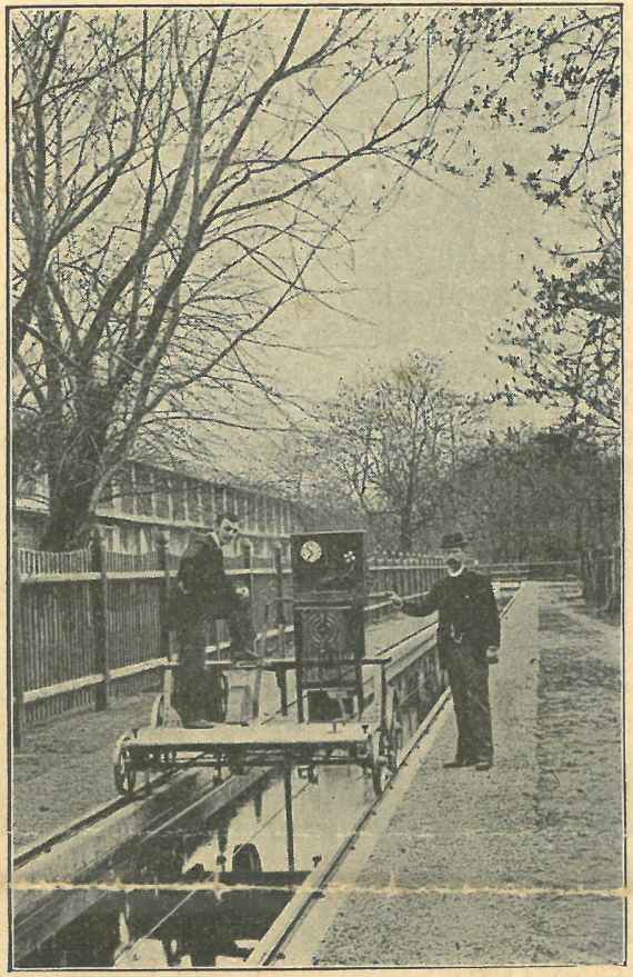 This picture shows an oblique view of the measuring trolley, which is standing on the measuring canal. There is an engine and a large box with a clock for recording measurement data on the measuring trolley, in front of it a rod immersed in water. On the far left of the measuring trolley is a worker, and on the right outside the current meters is another worker wearing a hat and pointing at the trolley. On the far left of the picture you can see a parallel house.
