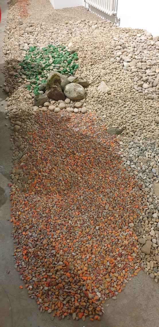 Along a wall, numerous stones lie on the ground and form an elongated gravel bank. The size of the stones ranges from 1 to 2 cm to about the size of a head. In one part of the gravel bank the stones are coloured red and in another part green.