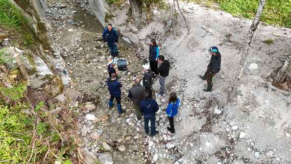 10 people are standing in a small stream with larger stones and little water. One person is holding a laptop and explaining something; the other people are listening.