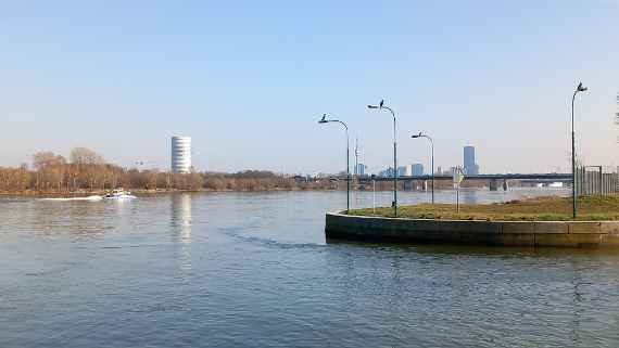 The picture shows the Danube in Vienna. In the foreground, you can see the entrance to the lock of the Danube Canal and the quay wall, on which four ducks are sitting. On the quay wall there are approximately five meters high waterstreet lighting, on each sitting a cormorant on top. In the left part of the picture is a speedboat, in the background, about four kilometres away, you can see the skyscrapers of Donaustadt in Vienna, as well as the North Bridge, about one kilometer away.