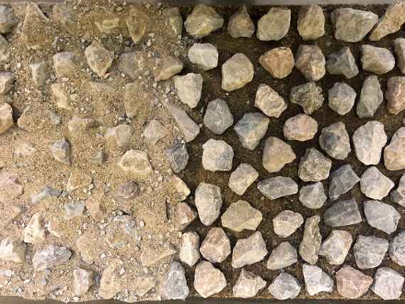 Photo shows stones about 5 cm in size lie loosely distributed on a flat surface. In the left half of the picture, the open areas between the stones are filled with sand.