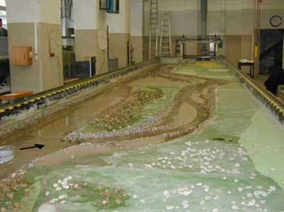 Physical model at mean water Q = forty-five cubic meter per second in a laboratory channel of the laboratory hall, the view is directed downstream, one can see two arms of water, on the left the bed load trap, on the right the bypass channel. The foreland areas are made of concrete in contrast to the water arms formed from sand.