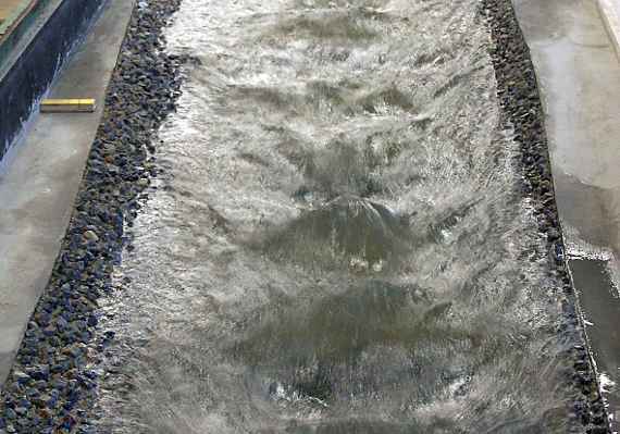 Photo of the model test. Water flows through a straight channel of about three meters width. The surface of the water shows high waves.