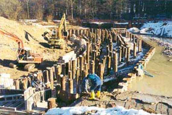 Erection of the wooden box weir in nature (foundations), you can see an abundance of wooden piles driven into the ground. Two excavators stand next to them. On them the wooden box weir will be built later. The river was diverted for the construction of the weir. It will be built in the dry.