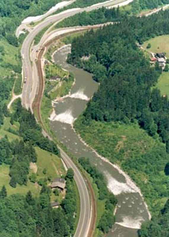 Aerial view of the project section on the Salzach after the rehabilitation, you can see an aerial view of the Salzach from the section in question. The river runs in a curvy course in the middle of the picture with the direction of flow from bottom to top. On the left side of the river runs the federal road and the railway line. On the right is a partly wooded, partly grass and bush covered river bank. The 4 newly built ramps for the stabilization of the riverbed are recognizable because of the white foaming water.