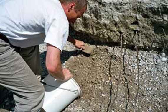 Removal of underlayer material from a renaturation section on the Isar river. Graduate engineer Schaipp pushes gravel from a river bank into a bucket with a shovel. The gravel layer is subdivided - in the upper part there is finer and more solidified material, interspersed with plant roots visible, whereas in the lower part there is coarser, loose gravel.