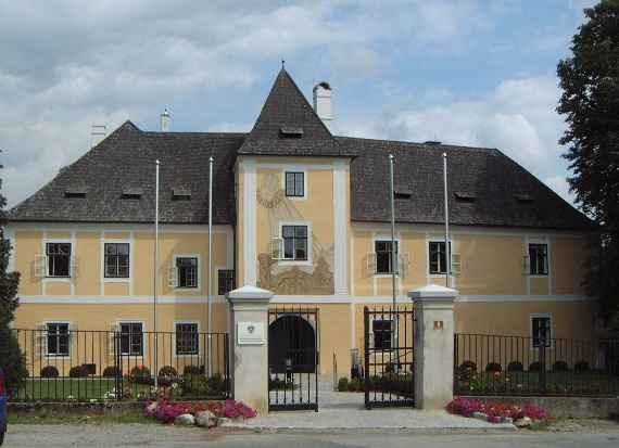 View of the building of the institute from the front, it is a castle in Petzenkirchen. In the foreground is a fence with gate. The gate is anchored on the left and right in two large columns. The building consists of a slightly higher central part (three floors high) and two side parts with two floors each).