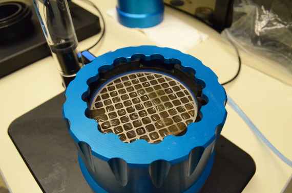 The photo shows a device for the so-called KSAT determination of a soil (KSAT is the acronym for a parameter describing the water conductivity of a soil completely saturated with water. The higher the water conductivity, the more water can flow through the soil in the same time). You see a black plate on a table and a blue plastic ring on it. In the ring is a soil sample on which a metal grid lies.