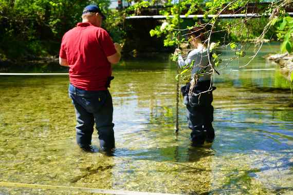Verschwommener Blick auf einen kleinen Fluss, in dem zwei Personen knietief im Wasser stehen und eine Messung durchführen.