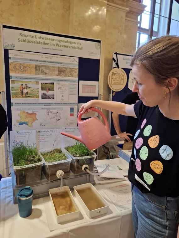 Person gießt Wasser mit rosa Gießkanne in transparenten Behältern mit Gras vor Poster "Smarte Entwässerungsanlagen als Schlüsselstellen im Wasserkreislauf".