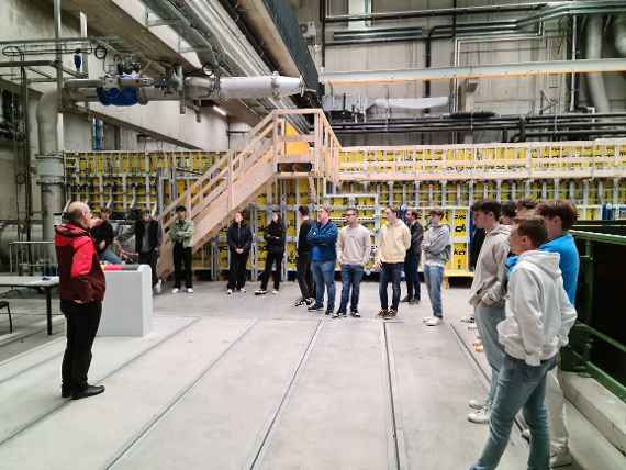 Viele Personen stehen in einer großen Halle des Wasserbaulabors und bekommen die Technik erklärt. Man sieht ein großes Rohr, dass aus dem Boden kommt sowie viele Kabeln und weitere Leitungen.