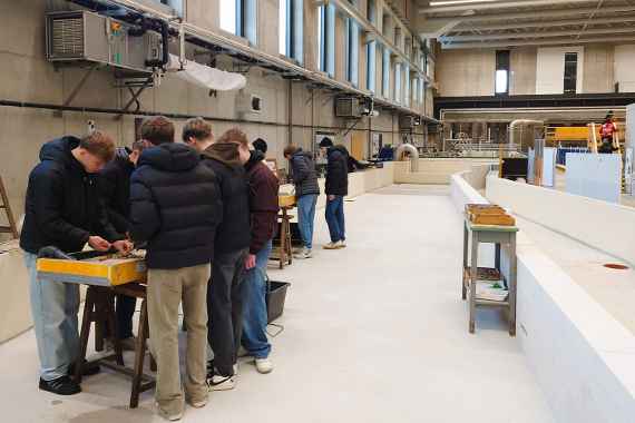 In einer großen Laborhalle sieht man Schüler an zwei Experimentierrinnen stehen, die gerade mit verschiedenen Materialien in diesen Experimentierrinnen bauen.