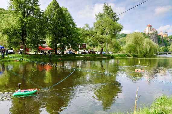 Blick vom Ufer auf einen Fluss, über den zwei Leinen gespannt sind, mit denen Messboote gezogen werden. Im Hintergrund auf einem Felsen das Schloss Vranov nad Dyjí.
