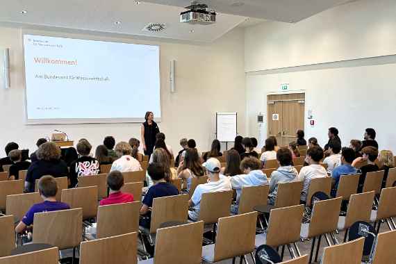 Ein großer Hörsaal mit Einzelbestuhlung, in dem ca. 40 Schüler und Schülerinnen in Richtung Leinwand und Vortragende schauen. Die Vortragende steht vor der Leinwand, auf dieser ist eine Willkommens-Folie projiziert.