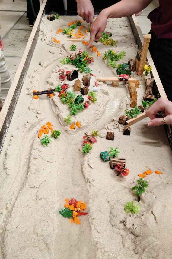 Vogelperspektive einer mit Sand gefüllten Experimentierrinne, in der Steine und Hölzer das durch mehrere Krümmungen fließende Wasser lenken.