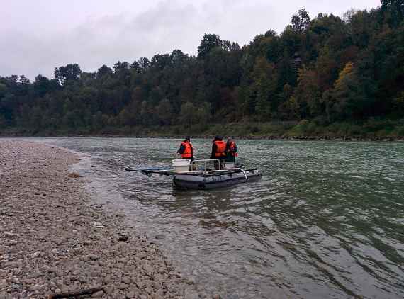 Drei Männer in einem Boot auf einem Fluss, die gerade Proben entnehmen. Links sieht man Land und rechts Wald.