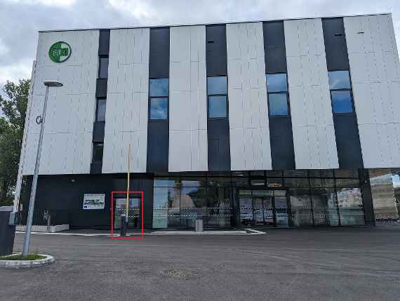 Das Hauptgebäude sieht man frontseitig. Links offene Schranke, daneben Lichtmast. Rot umrandeter Rahmen ist der derzeitige Eingang.