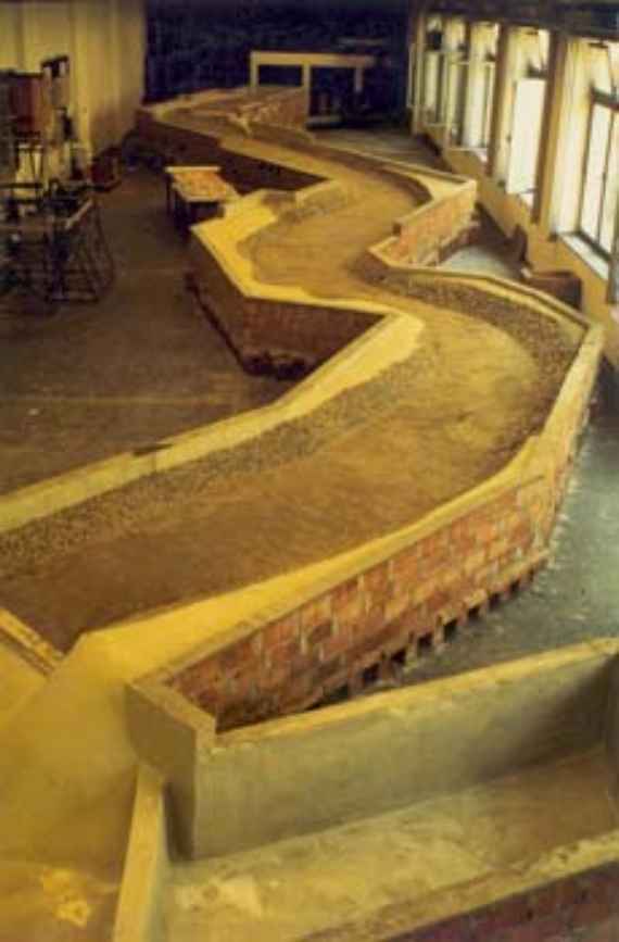 Salzach Eschenau im physikalischen Modell mit der Ausgangssohlenlage, man sieht das physikalische Modell im Überblick mit der Ausgangssohlenlage vor einem Experiment. Der geschwungene Gewässerlauf im mit Ziegel gemauerten Modellfluss führt durch die gesamte Laborhalle. Der Fluss hat ein Flussbett aus Sand und ein Flussufer, das zum Schutz mit Steinen belegt ist.